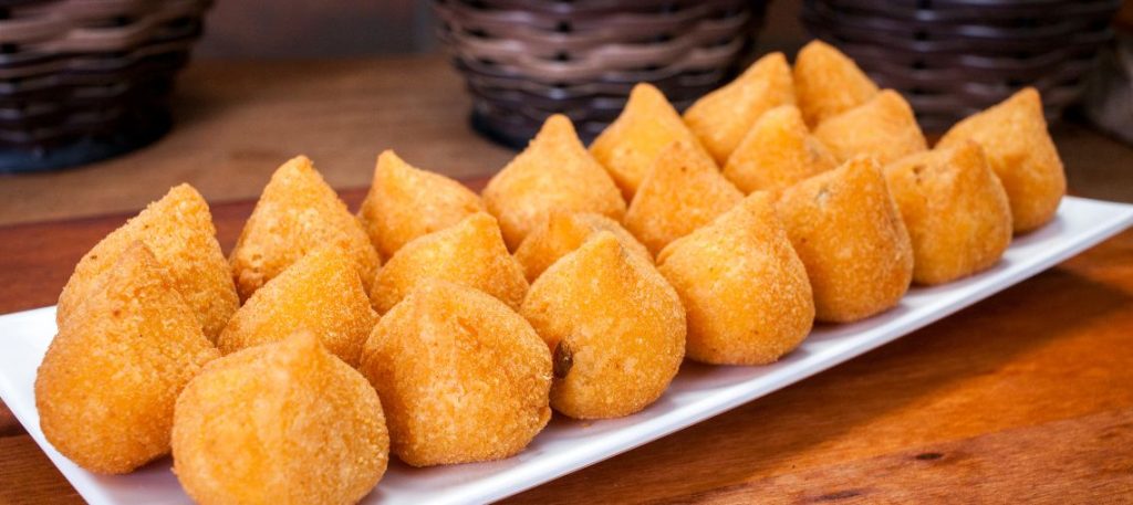 A coxinha de frango é um clássico da culinária brasileira, amada por muitos devido à sua casquinha crocante e recheio suculento de frango. Perfeita para festas, lanches ou qualquer ocasião, essa receita de coxinha de frango simples vai te conquistar pela praticidade e sabor. Ingredientes: Massa: 2 xícaras de farinha de trigo 2 xícaras de caldo de galinha (pode usar o caldo do cozimento do frango) 2 colheres de sopa de manteiga 1 pitada de sal Recheio: 500g de peito de frango cozido e desfiado 1 cebola pequena picada 2 dentes de alho picados 2 colheres de sopa de óleo ou azeite 2 colheres de sopa de salsinha picada Sal e pimenta a gosto Para empanar e fritar: 1 ovo batido Farinha de rosca Óleo para fritar Modo de Preparo: Preparar a Massa: Em uma panela grande, leve o caldo de galinha e a manteiga ao fogo até ferver. Adicione a farinha de trigo de uma só vez e mexa vigorosamente com uma colher de pau até que a massa desgrude do fundo da panela. Retire do fogo e deixe esfriar. Preparar o Recheio: Em uma frigideira, aqueça o óleo ou azeite e refogue a cebola e o alho até ficarem dourados. Adicione o frango desfiado, a salsinha, o sal e a pimenta. Misture bem e deixe cozinhar por alguns minutos para pegar sabor. Deixe esfriar. Montar as Coxinhas: Com as mãos untadas com um pouco de óleo, pegue pequenas porções de massa e abra na palma da mão. Coloque uma colher de recheio no centro e feche a massa, modelando em formato de coxinha. Repita o processo com toda a massa e recheio. Empanar e Fritar: Passe cada coxinha no ovo batido e depois na farinha de rosca, cobrindo bem. Em uma panela, aqueça o óleo para fritura. Frite as coxinhas aos poucos, até ficarem douradas e crocantes. Retire com uma escumadeira e coloque sobre papel absorvente para escorrer o excesso de óleo. Dica Final: Coxinha de Frango Simples